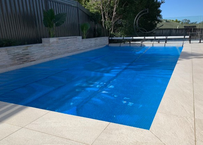 A swimming pool covered with a blue pool cover next to a tiled patio area. The pool, ensuring optimal pool safety, is surrounded by a fence and some plants in the background. The sky is clear and sunny. | AES Pool Heating Solutions