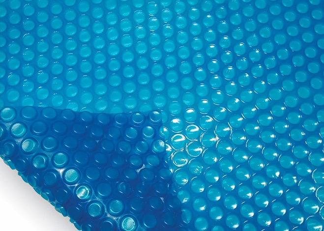A close-up image of a blue bubble wrap sheet, shiny and reflective, resembling pool covers. The bubbles are uniformly spaced and cover the entire surface. A portion of the sheet is lifted, revealing the bubbles' texture and translucence from underneath. | AES Pool Heating Solutions