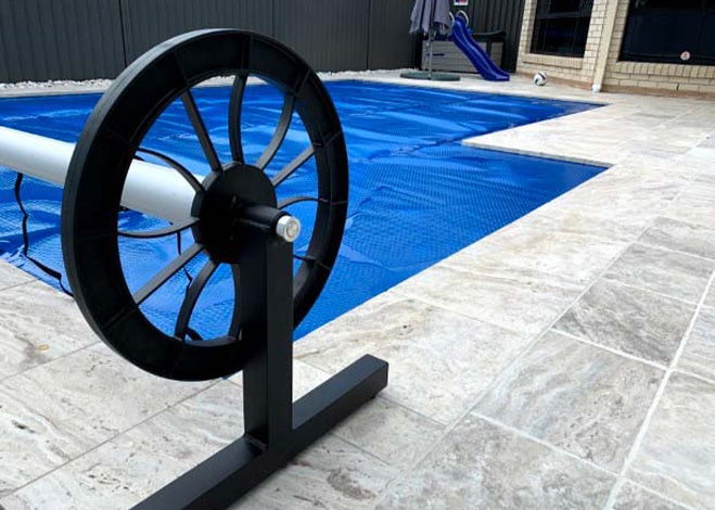 An outdoor swimming pool covered with a blue pool cover provides essential pool protection. A pool reel, partially seen in the foreground, is used to roll and unroll the cover. The area is surrounded by light-colored tile flooring, with a small slide and other structures visible in the background. | AES Pool Heating Solutions