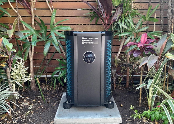 An outdoor heat pump by Australian Energy Systems is installed on a concrete slab in a garden. It is surrounded by various tropical plants and placed in front of a wooden fence. The heat pump, ideal for pool covers, features a digital display and black and blue color accents. | AES Pool Heating Solutions
